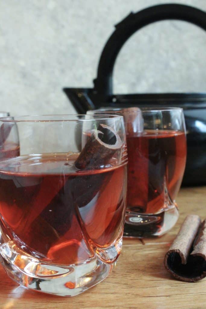 Two glasses of cinnamon tea with cinnamon sticks and a kettle behind it.