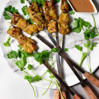 Chicken satay on a large plate with satay skewers and peanut sauce next to them.