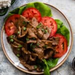 Beef stir fry on a plate over a bed of tomatoes and lettuce next to soy sauce and rice.