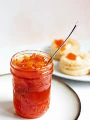 A jar of papaya jam with a spoon sticking out of the top with appetizers in the background.