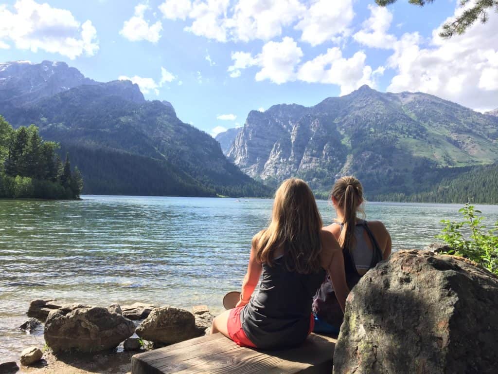 Two girls sitting and looking at the mountains next to a lake. 