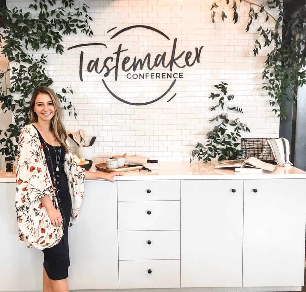 A girl posing in front of a kitchen set with a sign that says "Tastemaker Conference". 