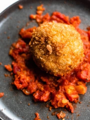 an arancini ball on a black plate sitting on a bed of tomato sauce.