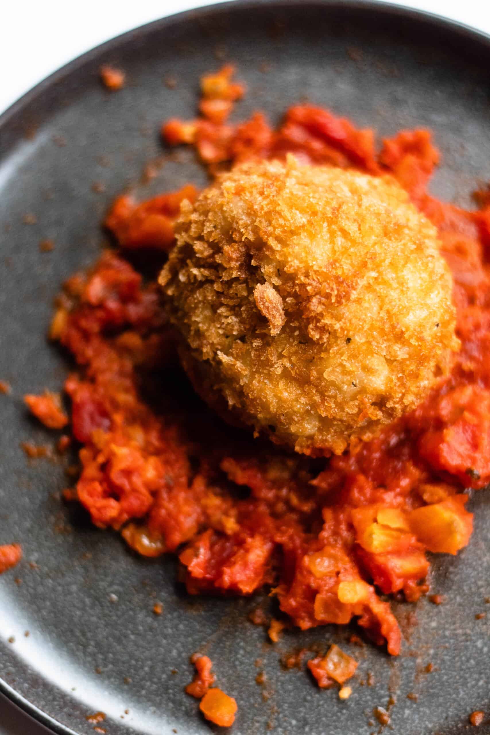 an arancini ball on a black plate sitting on a bed of tomato sauce.