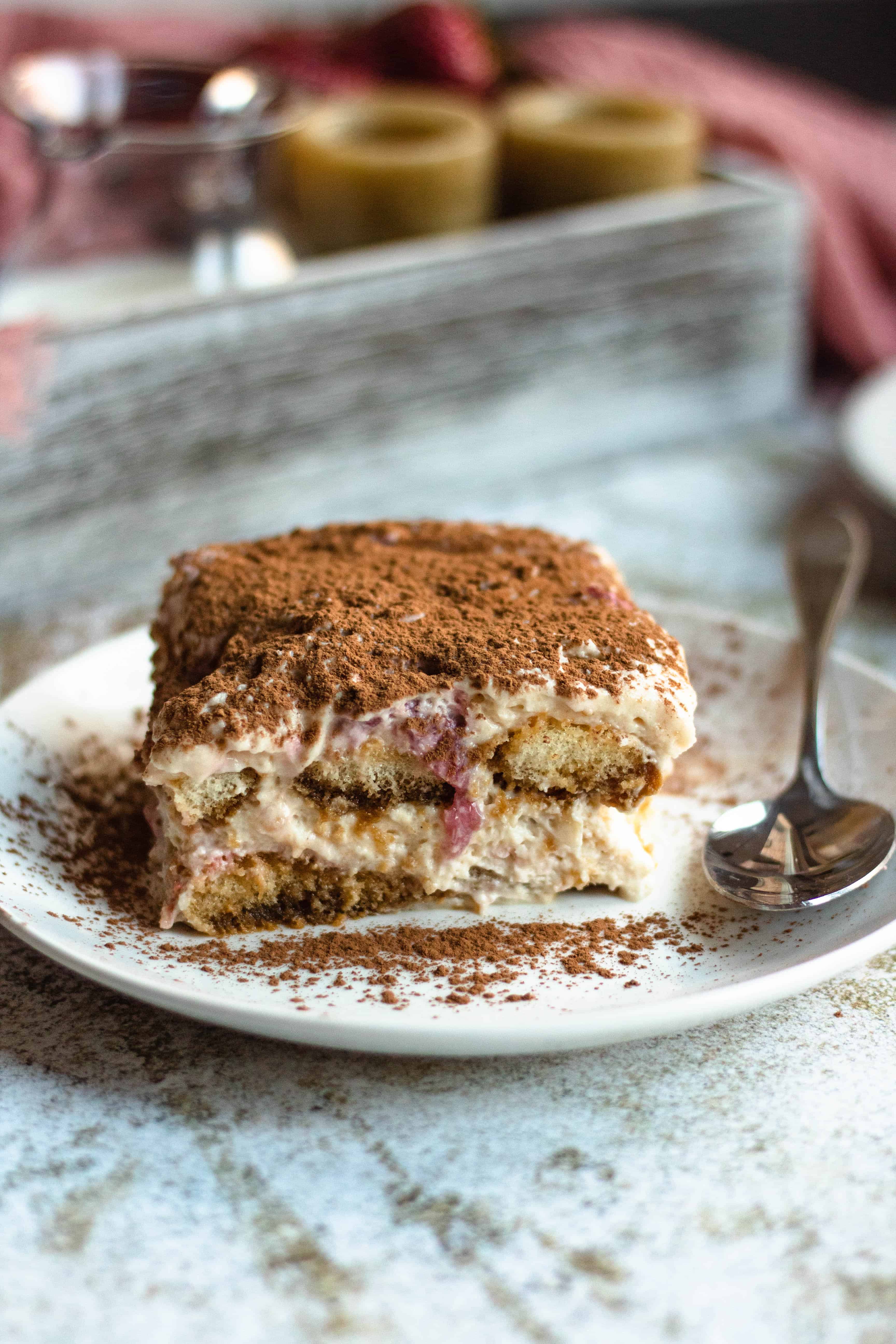 Slice of strawberry tiramisu, an Italian dessert recipe, dusted with cocoa powder on a plate with a spoon on the side.