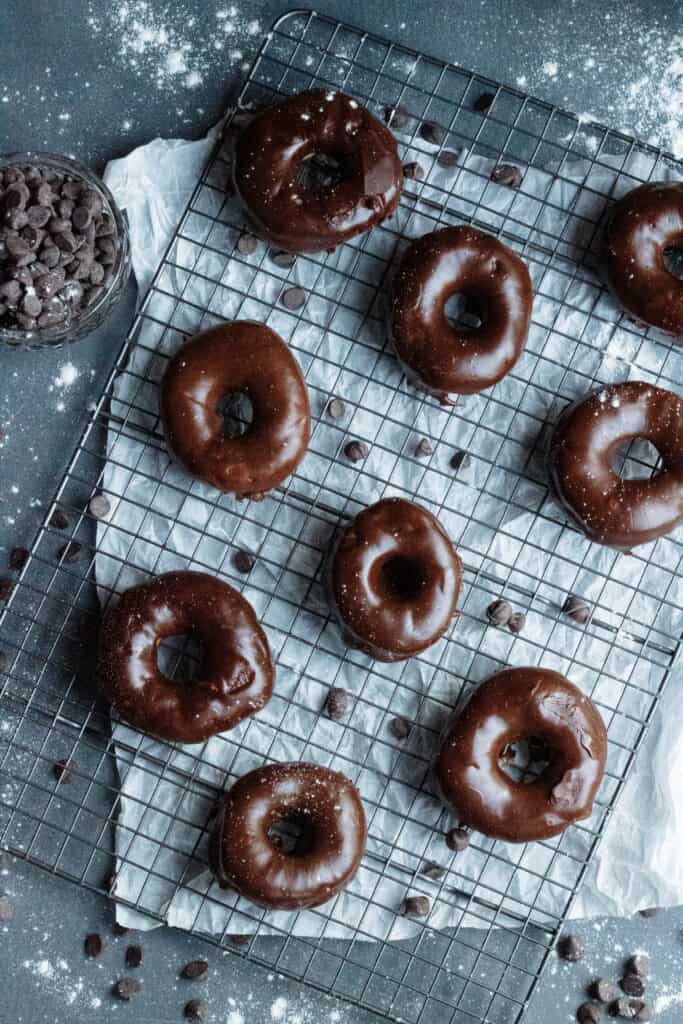 overhead view of chocolate glazed donuts on a cooling rack with chocolate chips and parchment paper