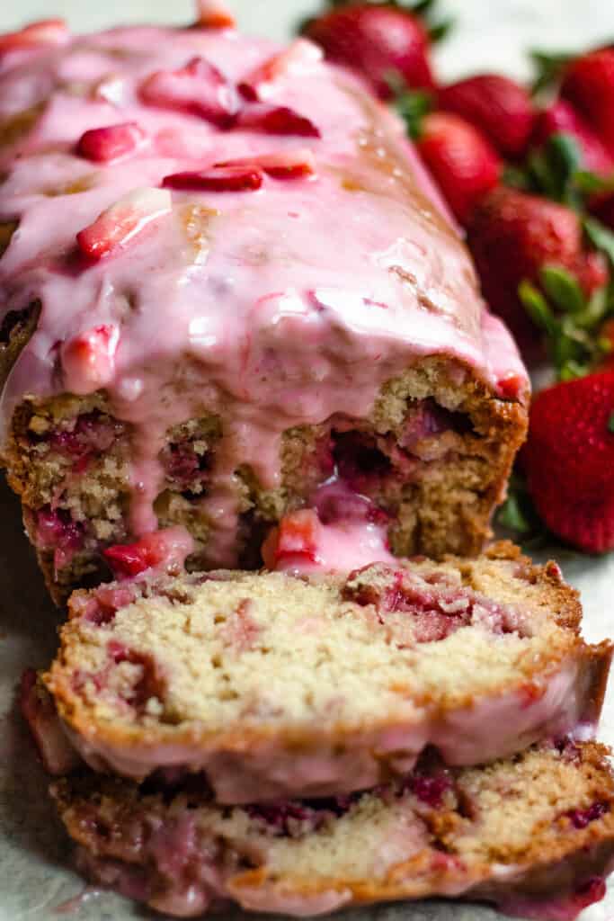 Sliced strawberry bread with glaze dripping down and strawberries in the background