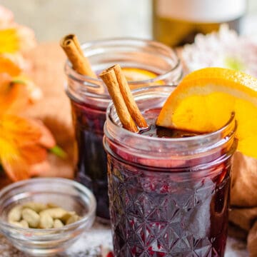 Mulled Wine in a glass jar with cinnam on sticks, oranges, and cardamom pods around it.