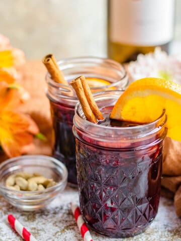 Mulled Wine in a glass jar with cinnam on sticks, oranges, and cardamom pods around it.