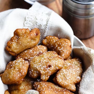 Banana fritters in a grey bowl with a lace linen sticking out of it, topped with powdered sugar.