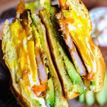 A hand holding a peameal bacon sandwich with fried egg and avocado.