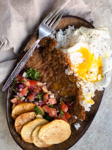 A plate of silpancho with slices of potatoes, salsa, cooked beef, white rice, and fried egg.