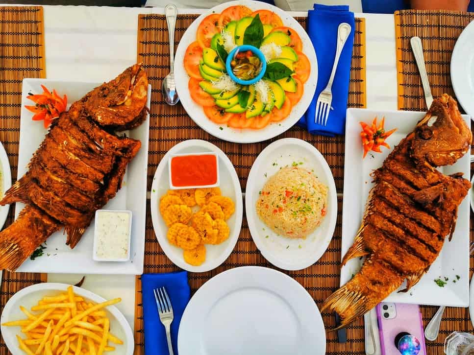 A table set with Dominican Republic food including cooked fish, rice, tostones, fries, and fruits and veggies. 