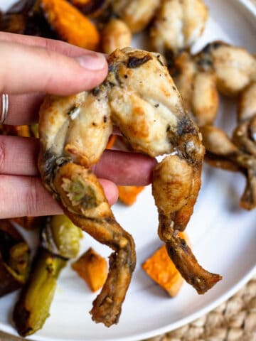 A hand holding frog legs with sweet potatoes in the background.