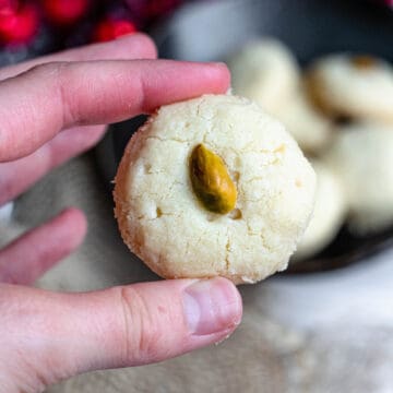A hand holding a butter cookie with a pistachio in the center.