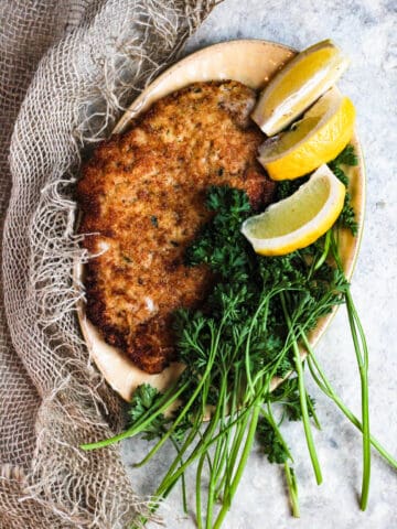 Wiener schnitzel on a plate with parsley and lemon slices surrounding it.