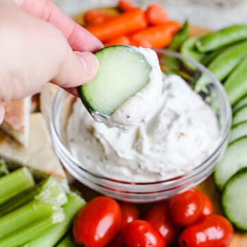 A hand dipping a cucumber into healthy veggie dip with cherry tomatoes and celery surrounding it.