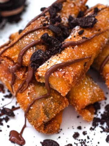 Oreo egg rolls in a pile with melted chocolate on top of them and crushed oreos surrounding them.