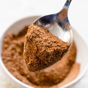 A spoon scooping up baharat seasoning from a small bowl.