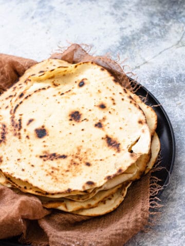 Cooked flour tortillas sitting on a large plate with brown linen beneath it.