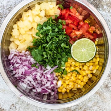 Pineapple corn salsa in a large mixing bowl with red onions, tomatoes, limes, and cilantro.