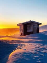 A wooden sauna in the winter, sitting in front of a sunrise.