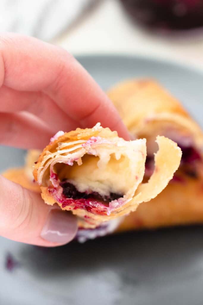A hand holding a Blueberry Cheesecake egg roll with a bite taken out of it.