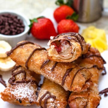 Banana split egg rolls piled on top of each other, topped with melted chocolate and powdered sugar and surrounded by fruit.