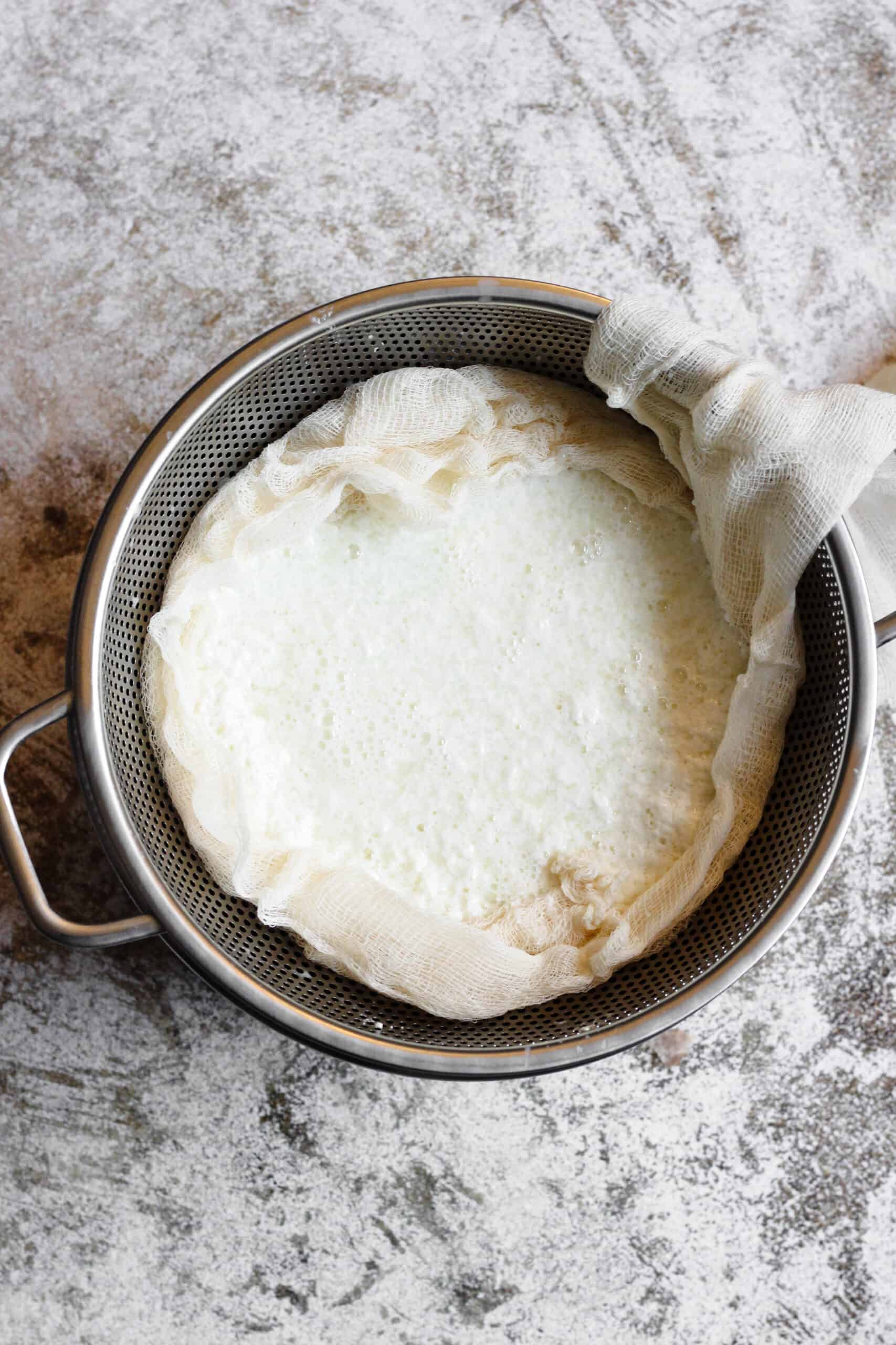 Homemade ricotta sitting in cheesecloth with a strainer below it. 
