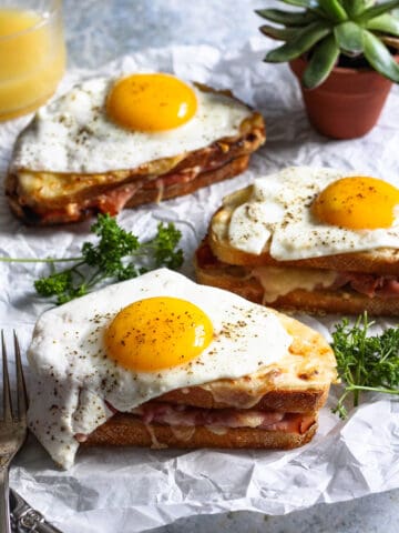 3 Croque Madame sandwiches sitting on parchment paper with parsley next to them.