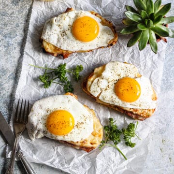 Croque madame sandwiches on parchment paper surrounded by succulents, parsley, and eating utensils.