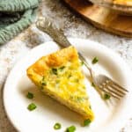 A slice of ham and cheese quiche on a plate with green onions and a fork.