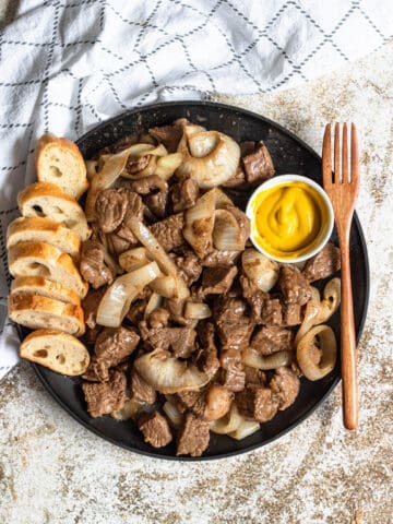 Cubed steak on a plate with cooked onions and baguette slices.