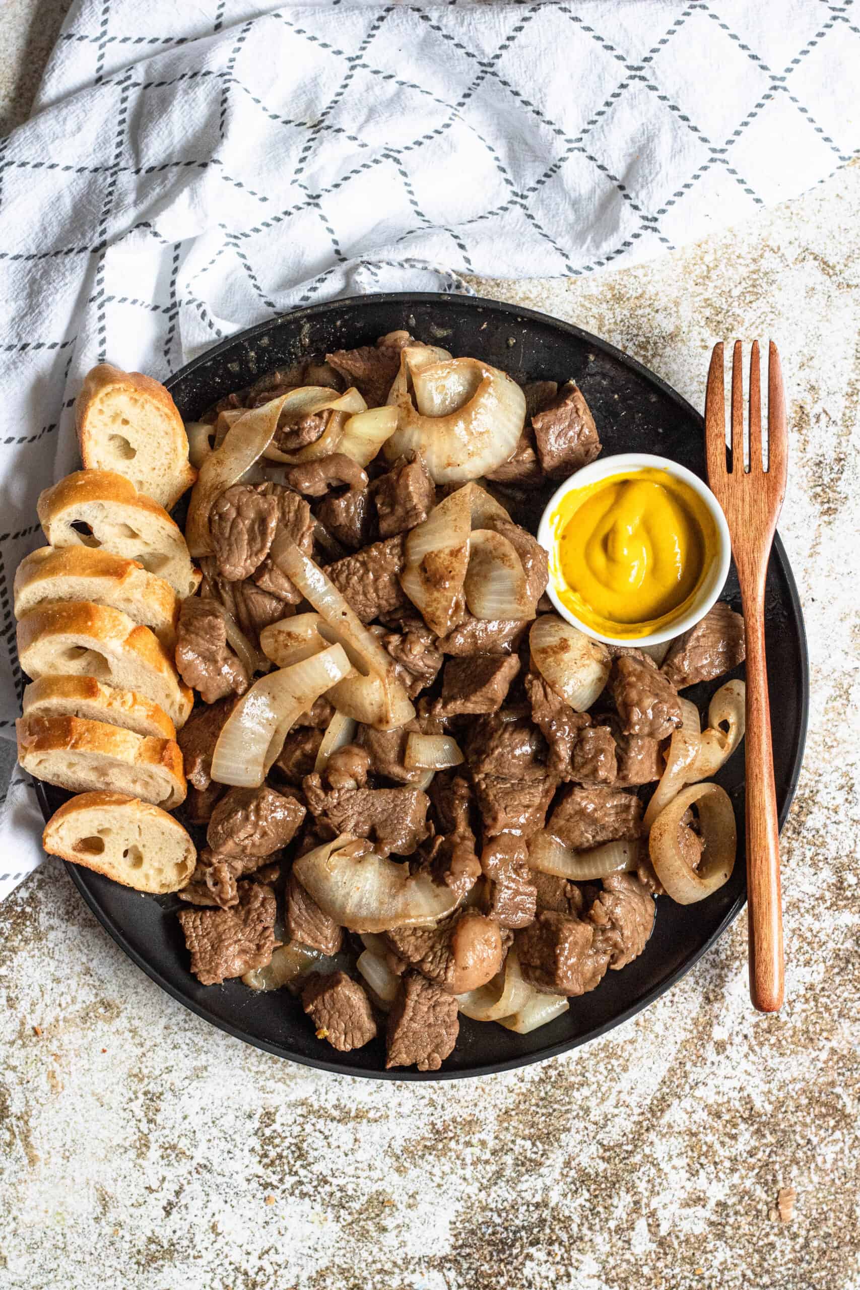 Cubed steak on a plate with cooked onions and baguette slices.