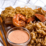 Pinterest image of a close-up shot of a small bowl of yum yum sauce with rice, noodles, and shrimp on a plate with chopsticks on the side.