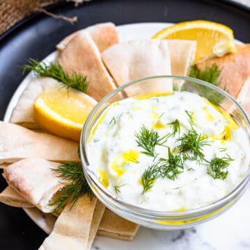 Tzatziki sauce in a small bowl, surrounded by pita bread and topped with olive oil and dill.