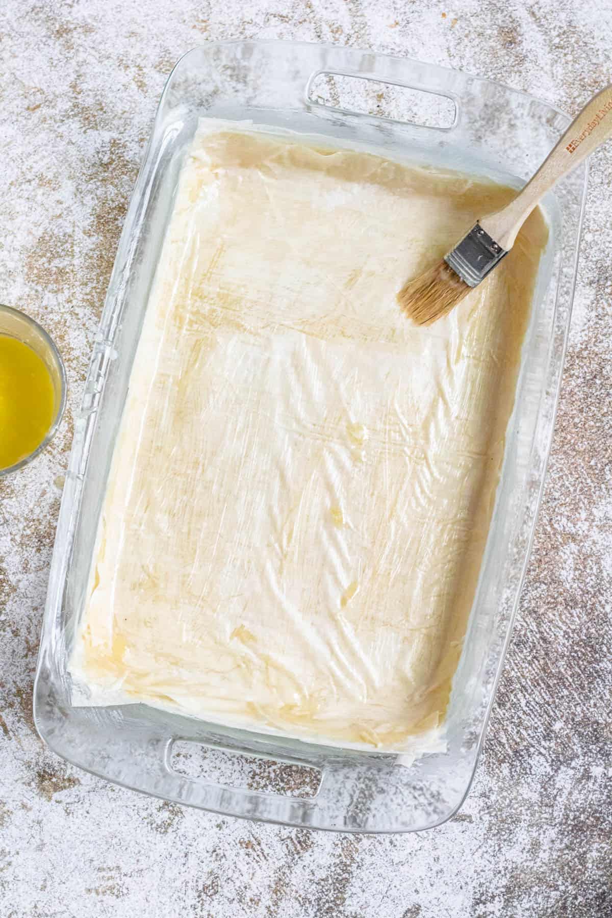 Phyllo dough being added to a baking dish and brushed with melted butter to prepare Pistachio Baklava. 