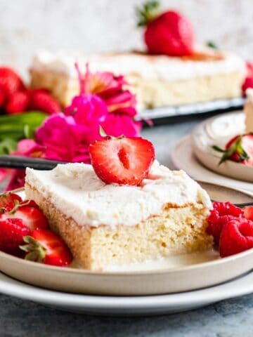 A slice of tres leches cake topped with strawberries and next to flowers.