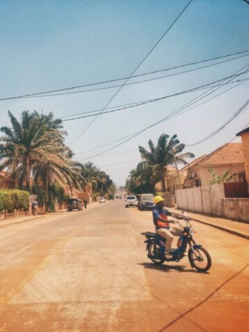 An orange road in Guinea Bissau with a man on a scooter in the road.