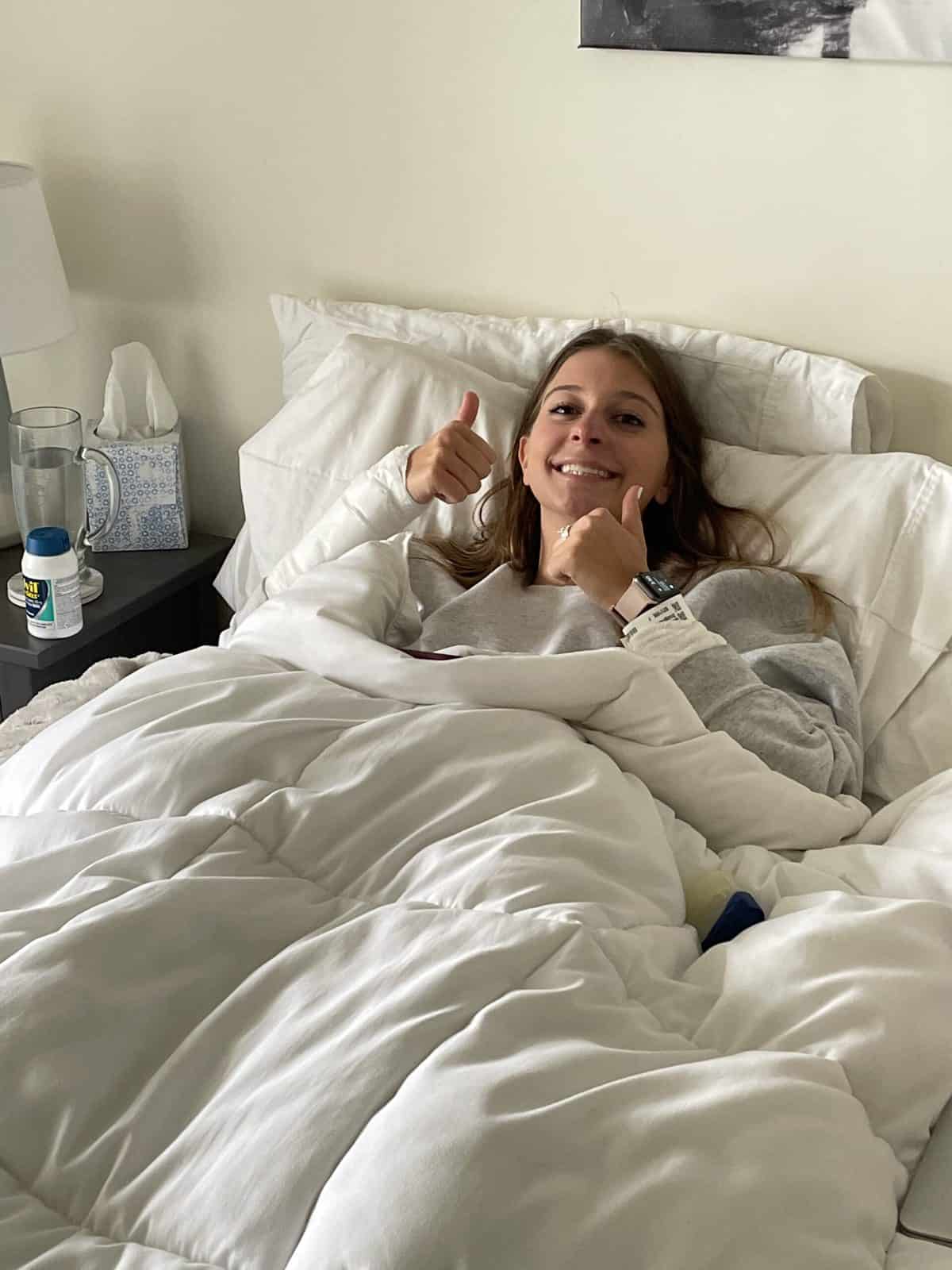 A woman laying in bed with a hospital bracelet on, giving a thumbs up sign.