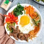 Beef Bibimbap in a large bowl serve with various side dishes and topped with an egg.