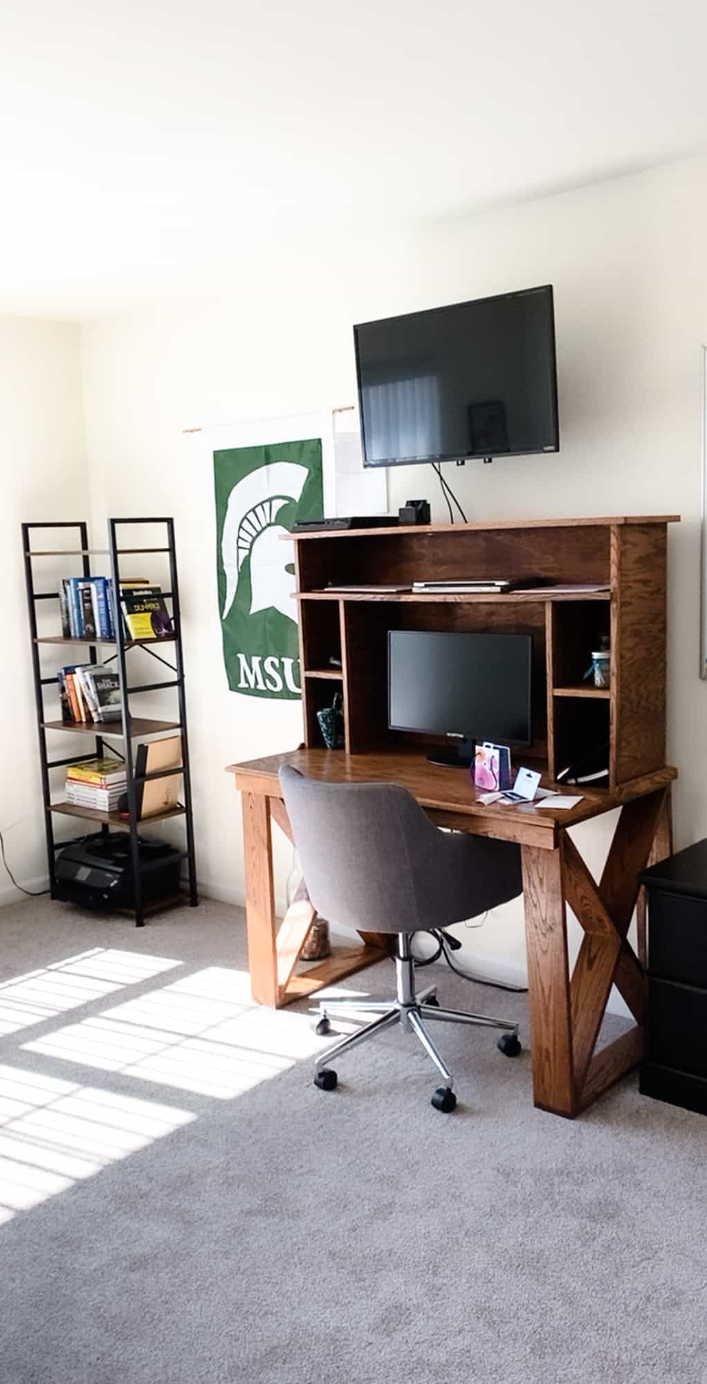 An office with a desk, a television, a flag, a bookshelf, and a computer.