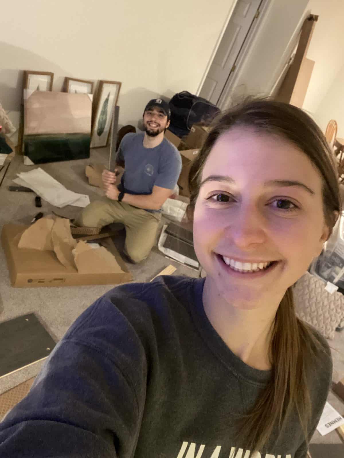 A girl taking a selfie with a man in the background assembling furniture.