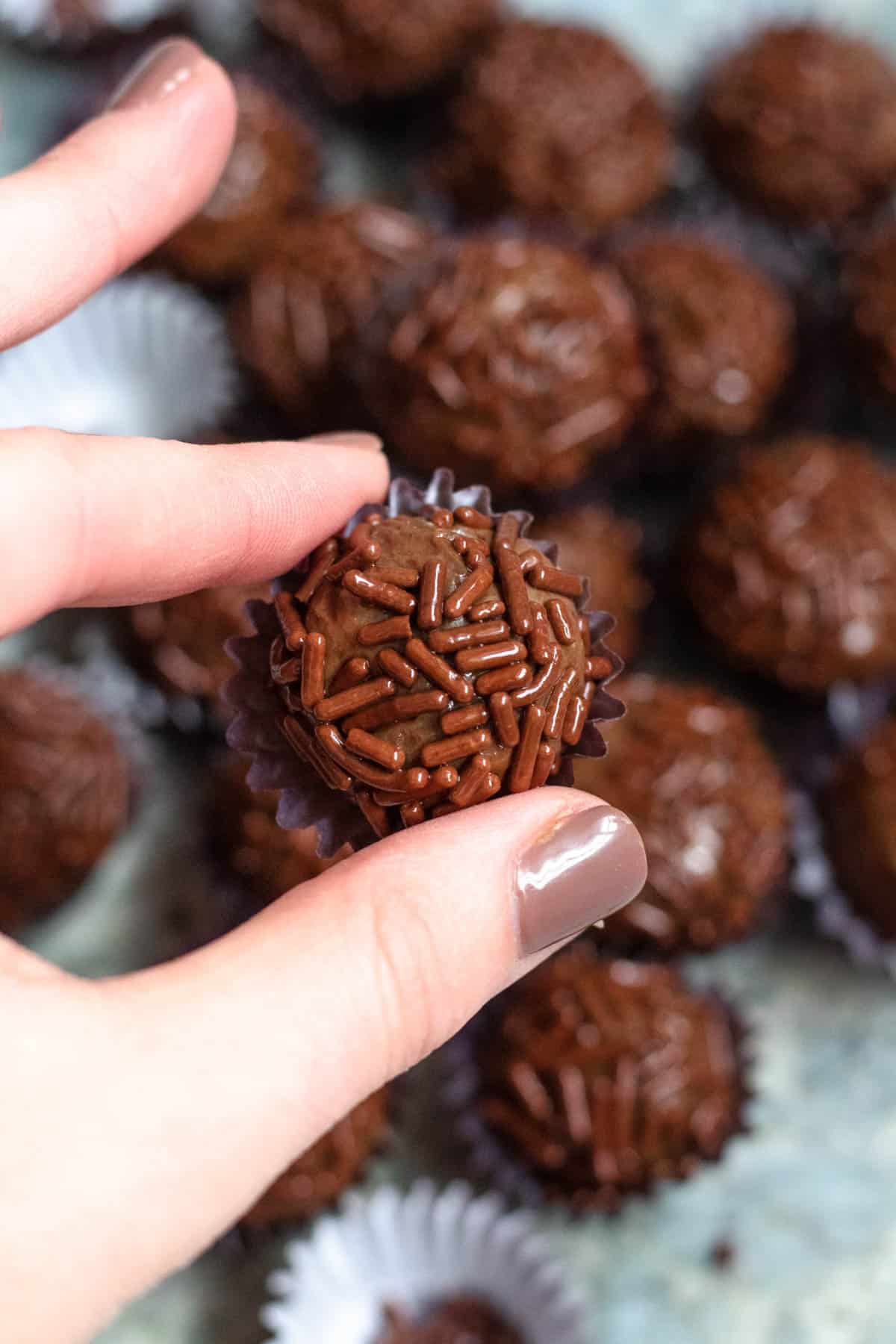 Fingers holding up a single Brigadeiro in front of the batch of them. 