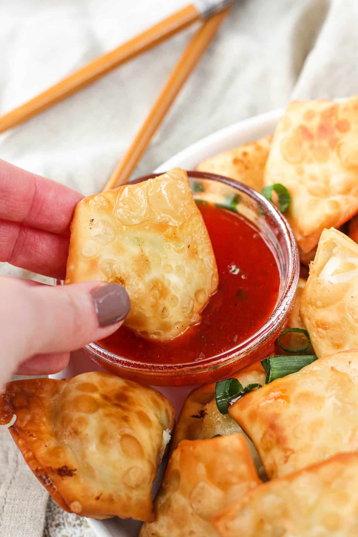 Hand picking up a crab rangoon and about to dip into the red sauce. 