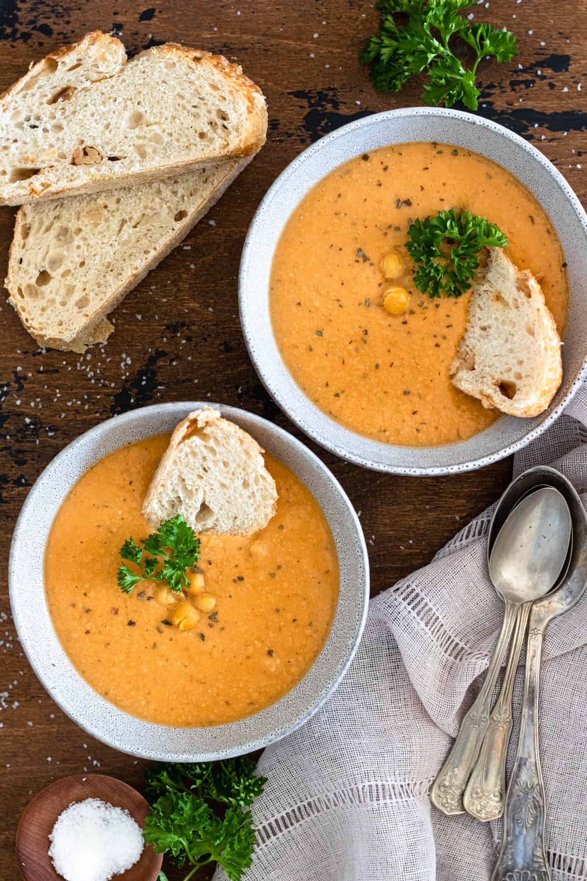 Top view of two bowls of soup served with whole chickpeas on top as garnish and slices of bread on the table next to them. 