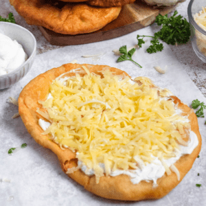 A Hungarian flat bread topped with sour cream and sprinkled with shredded cheese in front of parsley and garlic.