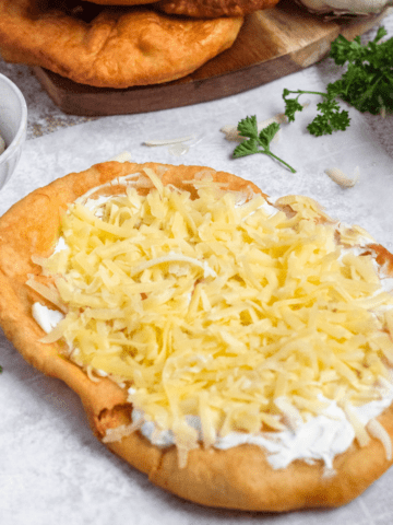 A Hungarian flat bread topped with sour cream and sprinkled with shredded cheese in front of parsley and garlic.