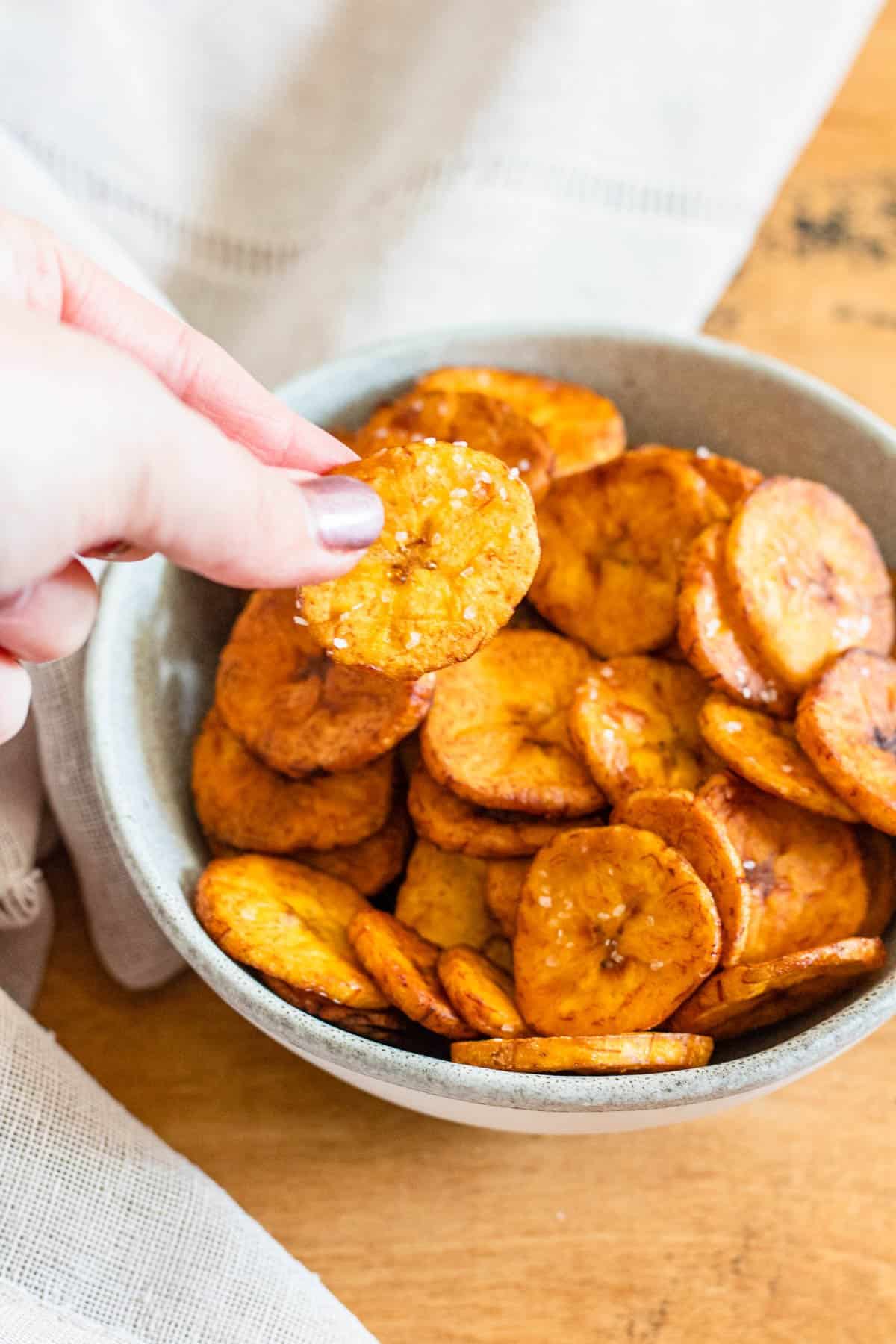 Fingers picking up a sweet plantain chip from a bowl. 