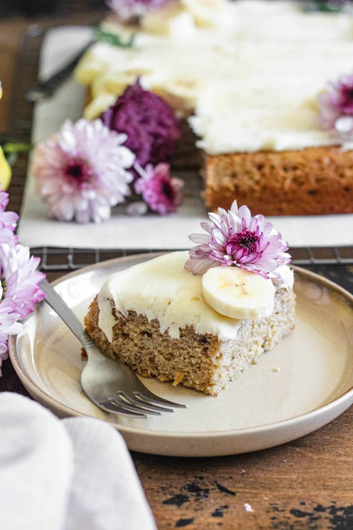 Slice of banana cake garnished with banana slice and a small purple flower sitting on a plate with a fork. 